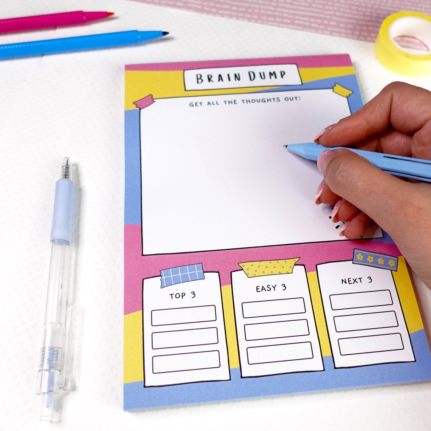 Person writing on an ADHD A5 'Brain Dump' notepad with stationery items on a white surface