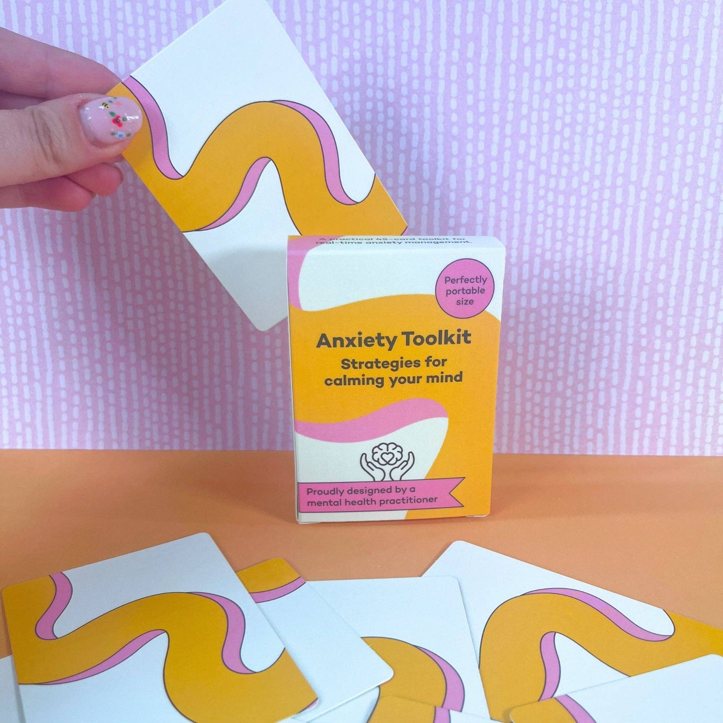 Hand reaching for a card from the ADHD mental health anxiety toolkit with cards on a pink and orange background