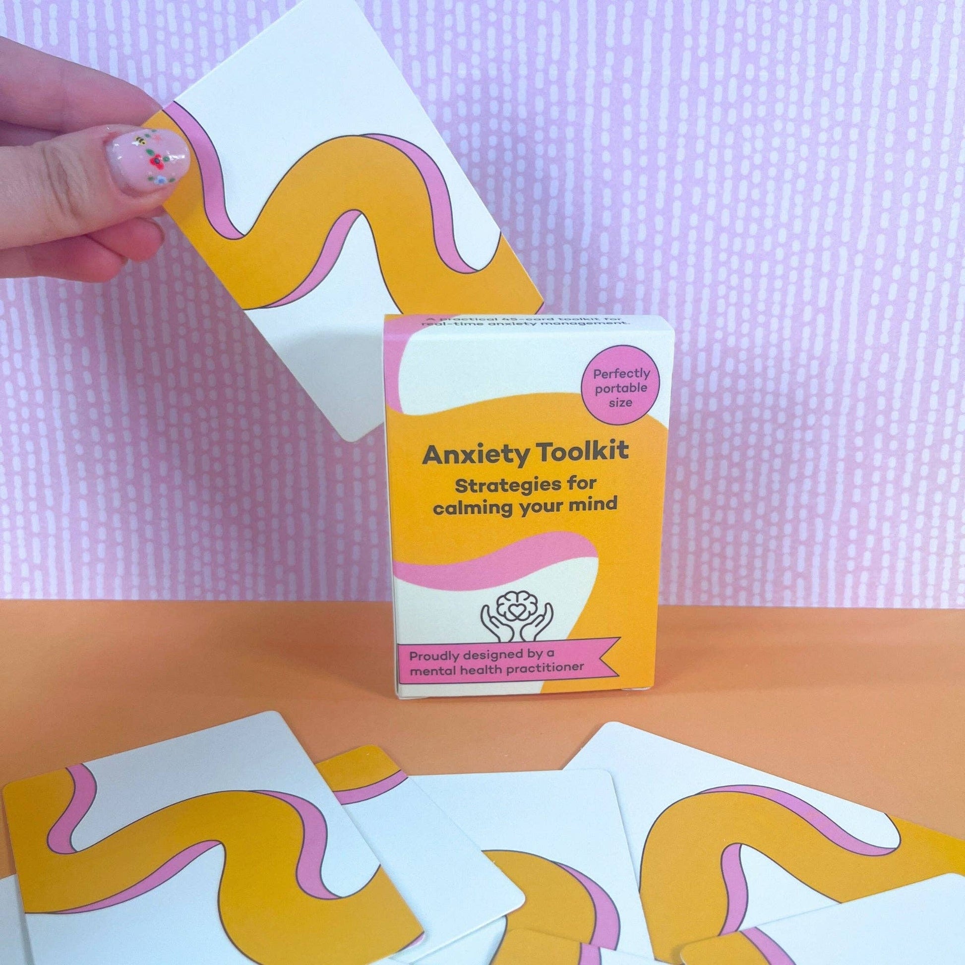Hand reaching for a card from the ADHD mental health anxiety toolkit with cards on a pink and orange background