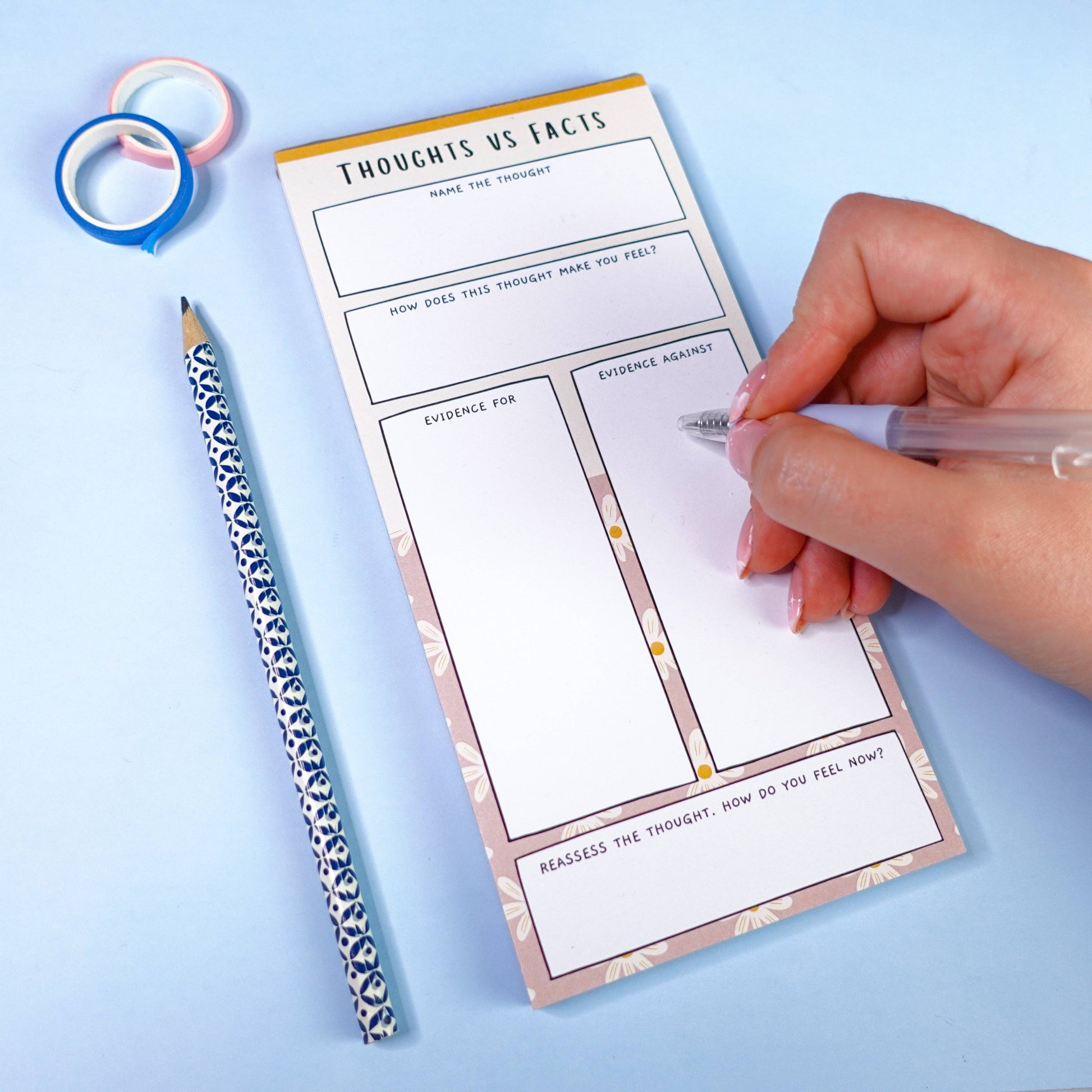 Hand holding a pen over a 'Thoughts vs Facts' ADHD notepad on a light blue background, next to a patterned notepad and coloured washi tape